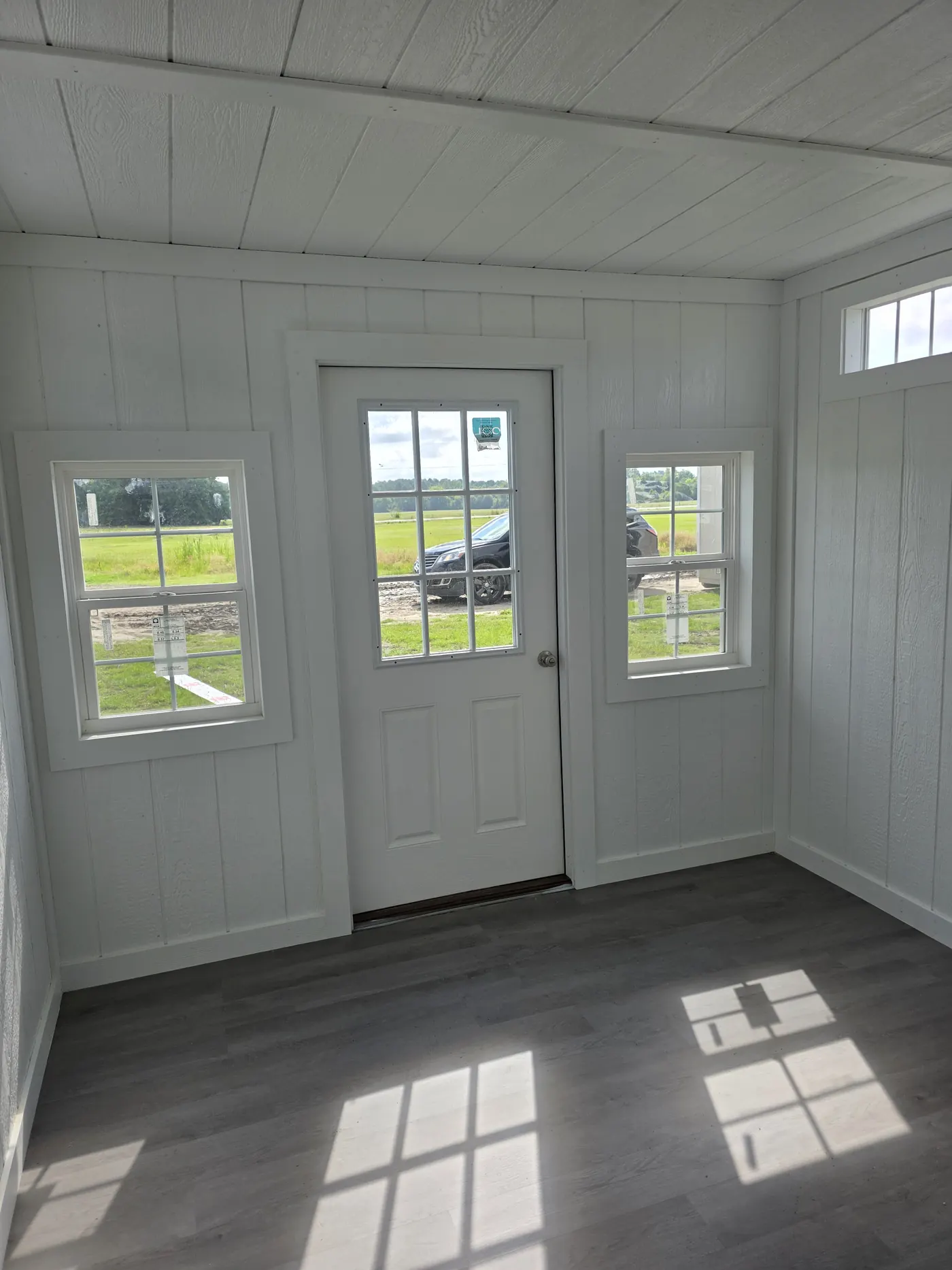 Finished shed interior with LVP flooring, white wall sheathing and a nine-lite entry door between two windows