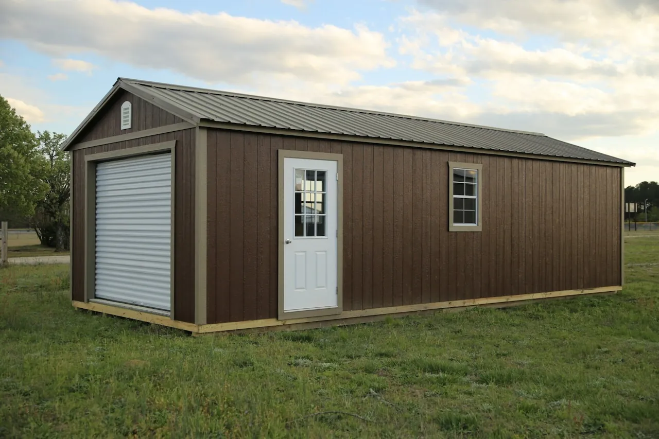 Brown lofted workshop shed with a white roll-up door, entry door and two windows on a dealer lot